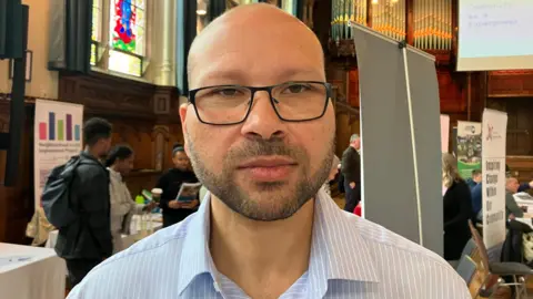 Twenty-nine arrests over recent violence Sameh Hassan, with short hair and black glasses in a blue striped shirt. Behind him is a crowded room in the Guildhall in Londonderry.