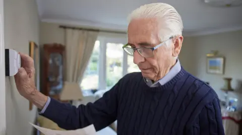 What is the winter fuel payment, how much is it and who gets it? Getty Images An elderly gentleman wears a blue jumper in his living room at home. He has an energy bill in one hand and is adjusting the thermostat with the other.
