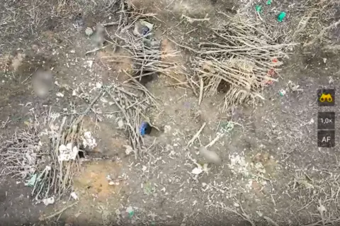 A Russian soldier takes the stand for an execution The three frontline foxholes dug in the ground and covered with sticks as seen by a Ukrainian drone filming shortly after the operation.
