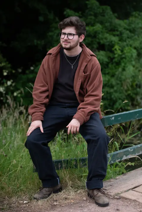 A preacher asked me out and then started stalking me A man in a brown jacket with black trousers sits with a long green meadow and bushes in the background. He is sitting on a green wooden gate.