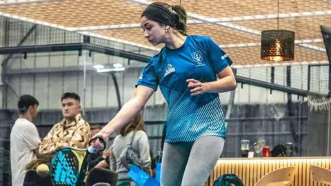 'I want to play the sport but I can't afford it' Daisy Whitlie A young woman wearing a blue sports team top and grey leggings swings low at an incoming ball. Spectators mill around in the background of the shot, which appears to have been taken inside a leisure centre with high concrete walls.