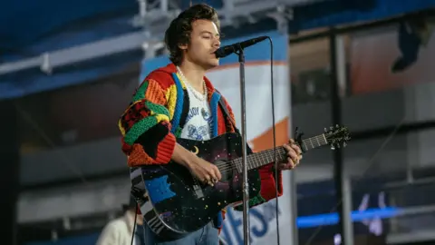 NI-born designer Jonathan Anderson becomes fashion brand's creative director Getty Images Harry Styles stands on a stage holding a black guitar with moon and star details while singing into a microphone. He has longer dark brown hair and is wearing a long pearl necklace, a red, yellow, green and blue patchwork crochet cardigan and a white t-shirt underneath.