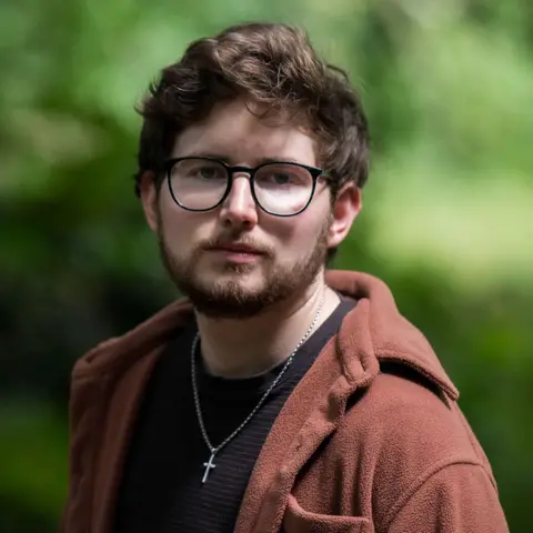 A preacher asked me out and then started stalking me A man in a brown jumper with a black T-shirt stands wearing glasses in front of a green bush with a cross necklace.