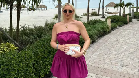 Asos customers banned over returns say it is 'slap in the face' A woman with blonde hair wears a strapless magenta dress, sunglasses and gold necklace and bangles. She holds a white Louis Vuitton purse and is smiling against a backdrop of white sand, palm tree and green shrubs.