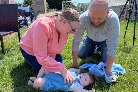 Parents' worldwide search for babies with son's rare condition Family photo Amanda and Nick are kneeling on grass playing with Jack. Amanda has a pink long-sleeved top, wears glasses and has her fair hair tied back in a pony tail. Nick has a beard and is wearing a grey jumper and jeans. Jack is in a blue baby grow and has a feeding tube going up his nose.
