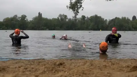 Failing water sector needs stronger regulation, review says PA Media Five swimmers in black wet suits and coloured swim caps in water behind a sandy beach. There are trees in the background.