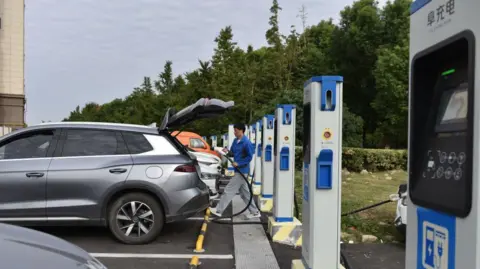 How China made electric vehicles mainstream Getty Images A man charges an electric car at an electric vehicle charging station on a street in Fuyang, China. Cars are parked in front of a row of blue and white charging stations.