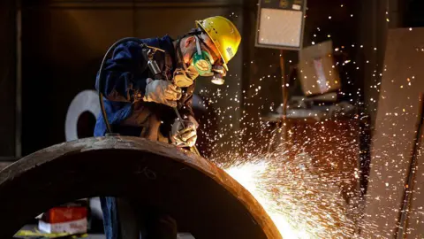 Turmoil and trade wars dominate 'summer Davos' Getty Images A person wearing a bright yellow helmet, goggles and a mask covering their lower face works in a factory amid a shower of sparks