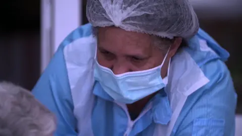 Covid inquiry begins examination of impact on social care sector BBC A care worker in a face mask and other personal protective equipment leans towards a care home resident. This shot was taken in April 2020.