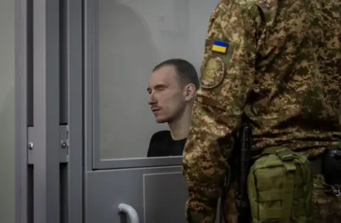 A Russian soldier takes the stand for an execution Dmitriy Kurashov, who has short hair and is wearing a dark top, sits in the dock watched over by a Ukrainian guard.