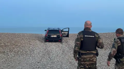 Smugglers deploy 'taxi boats' to collect migrants off the French coast Marianne Baisnée/BBC Two police officers walk towards an abandoned black car sitting on a shingled beach, one door still open