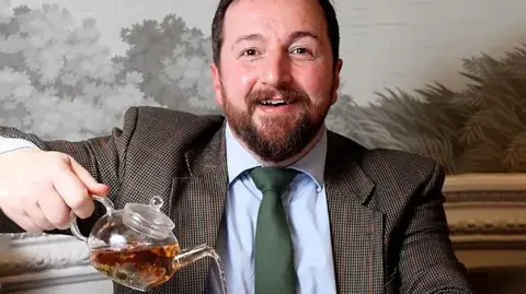 Fraudster jailed for selling fake 'Scottish-grown tea' Rex/Shutterstock Thomas Robinson wears a suit and pours tea from a glass tea pot into a white cup and saucer. He has short, brown hair and a short, brown beard and is smiling at the camera.
