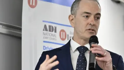 Learners struggle to pay before test, but instructors say pricing fair Pro Vision Photography Ltd A man with short grey hair in a dark suit, white shirt and navy tie stands on stage, speaking into a microphone