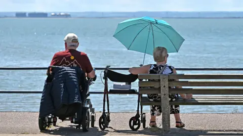 Heatwave peak set to bring 34C to parts of UK Getty Images A man and woman sit in the sun overlooking the sea. She shelters under a blue umbrella.