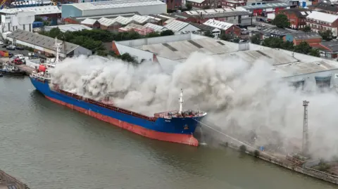 Ship on fire at Hull's Albert Dock as roads and shops are closed Dale Baxter/BBC An aerial view of a blue and red vessel with grey smoke billowing from the deck. There are industrial buildings in the background.
