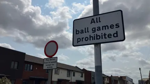 Ban 'no ball games' signs to get children off devices, play experts say Vanessa Clarke/BBC Two signs are on a residential street lined with houses. One reads 'All ball games are prohibited'. The other says 'Play Street 8am to sunset except for access'.