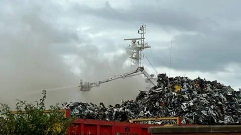 Ship on fire at Hull's Albert Dock as roads and shops are closed Becki Bowden/BBC A white aerial platform shoots water towards a fire on a ship. A pile of scrap metal is in the foreground.