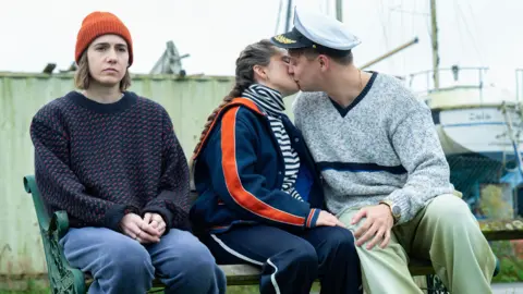 The dark comedy inspired by 'bleak' dating scene BBC/Various Artists Limited/Vishal Sharma From left to right: Sitting on a bench Josie (Sadler) is wearing blue trousers and a blue jumper with an orange beanie. She is sitting next to Billie (Davidson) wearing a blue tracksuit with a striped top and is kissing Nicky (Sam Buchanan) who is wearing green trousers a grey jumper and a sea captain's hat.