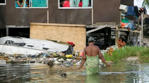 One in three citizens apply for climate change visa Getty Images A woman wrapped in a green patterned sarong wades through knee-deep water to reach her home. The house is on stilts and surrounded by rubbish. Washing hangs off the side of the building, and a small tin dinghy is upside-down beside the building.