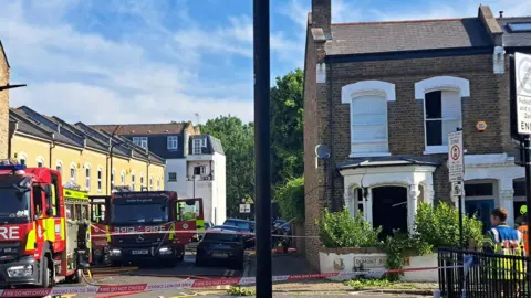 Murder arrest after woman stabbed PA Media A house with a blown out window on a residential street, with fire engines parked along the road