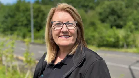 Government jobs support plans 'a mess', DWP officials tell BBC Karen Jones, who runs GoodLives charity in Cumbria, is pictured smiling. She has brown hair with blonde highlights and is wearing glasses which have dark frames. She is wearing a black work uniform which displays the charity's logo.