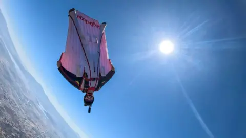 Scottish wingsuit flyer dies during Swiss Alps jump Byrne family Man - Liam Byrne - wearing wingsuit, upside down in mid air and smiling at camera, with sun in sky.