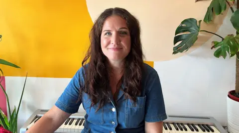 Why is Bristol's birth rate falling faster than other UK cities? Amy Joce smiles at the camera as she sits in front of an electric keyboard and some plants. She is wearing a denim jumpsuit and has long brown hair.
