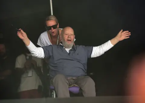 Old school charm from another era PA Media Michael and Emily Eavis going on to the Glastonbury stage - Michael Eavis is using a wheelchair and is wearing a blue top with a long-sleeved white top underneath and has his arms open wide