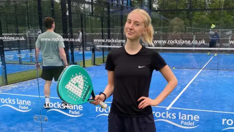 'I want to play the sport but I can't afford it' Rosie Hepworth Rosie is standing in front of a blue astro padel court. She is wearing a black t-shite and matching tracksuit bottoms and we can only see her from the waist up. She is holding a turquoise padel racket in one hand and the other hand is on her hip. Rosie has shot blonde hair, which has been tied back into a low ponytail. She is smiling into the camera with no teeth. Behind her are two men playing on the court, who can be seen through the glass walls. There is white branding written on the glass.