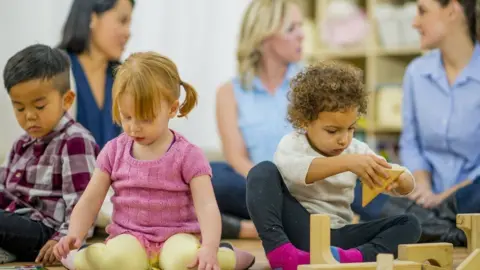 How can working parents get 15 and 30 hours of free childcare? Getty Images Three young children sit on the floor playing with toys in a library, with their mothers sitting behind.