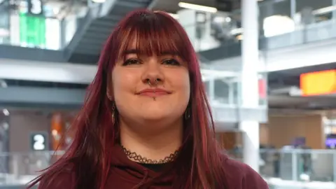 Video games cost a whole day's wages say fans, as prices rise BBC A woman with long brown hair, wearing a maroon shirt and a necklace, smiles at the camera