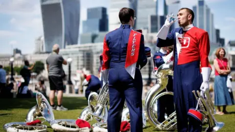 Health alerts come into force ahead of second heatwave EPA/Shutterstock Two members of the Pride of Dayton Marching Band drink water as they take a break from performing at Potters Field Park during a hot day in London. They are dressed in band uniforms of blue, white and red. They stand against a background of brass instruments lying on grass.