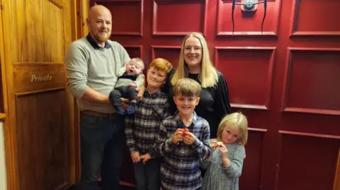 Parents' worldwide search for babies with son's rare condition Family photo Nick and Amanda with their four children, three boys and a girl. The boys are wearing shirts and the girl wears a dress, and all are smiling in front of a red panelled wall.