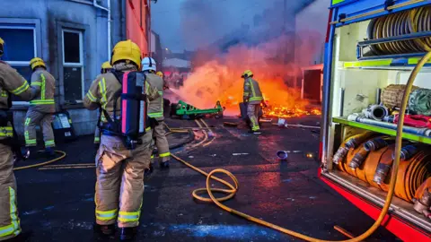 Disorder after alleged sex assault 'unwarranted', says Jim Allister Pacemaker Firefighters in brown suits and yellow helmets on the street holding a hose. There is a fire engine to the right and houses in front. There is a fire on the street which is being tackled by one of the firefighters. To the right of the image is the side of a fire engine with hose reels visible.