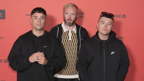 Kneecap performing at Glastonbury 'not appropriate', PM says PA Media Kneecap members Moglai Bap, Mo Chara, DJ Provai standing together in front of a red wall.
