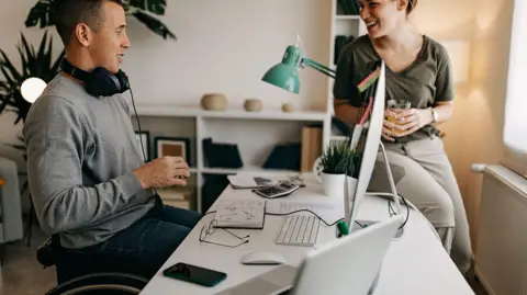 What are the Pip and universal credit changes and who will be affected? Getty A young man in a wheelchair who is working at a computer in the office talks to a female colleague who perches on the corner of his desk.