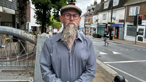 NHS and education named as local priorities A man with a long grey beard, wearing a blue shirt and a brown cap and glasses. He is looking directly at the camera. Behind him to the right is a road and traffic lights. To the left of him is the high street and a metal bench.