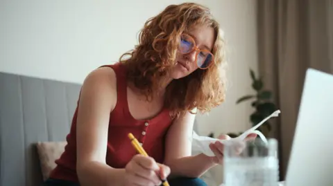 Lifetime ISAs leave some with less money than they put in, MPs warn Getty Images Young woman with curly hair and glasses sits at her laptop, making notes while looking at bills