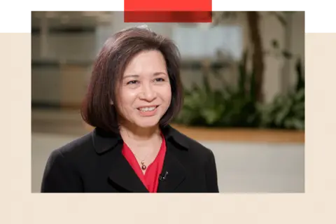 The US factory that lays bare the contradiction in Trump's policy A woman, wearing a black jacket, sits inside a room, smiling.