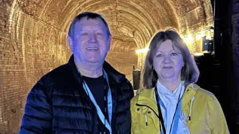 How is your NHS hospital doing on waiting times? John Winnik John Winnik and Lyn Williams are standing in front of a lit tunnel. He is in a dark puffer jacket and she is in a yellow rain coat.