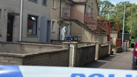 Second arrest in Donaghadee murder inquiry A forensic investigator standing outside a row of terraced properties at Elmfield Walk in Donaghadee where Sarah Montgomery died on Saturday. The investigator is dressed in a white hazmat suit and is holding a clipboard. The properties are two stories in height and there are a couple of sets of concrete steps leading to the first floor properties. There are cordons to the front and side of one ground floor property.