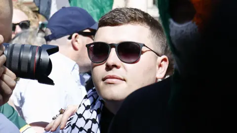 Rapper released on unconditional bail over terror charge EPA A close-up of a man in the midst of a crowd. He is wearing sunglasses and a black and white keffiyeh headdress around his neck like a scarf. There is a camera lens being held close to his face as a photographer takes a photo