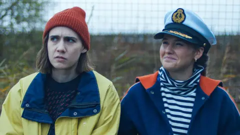 The dark comedy inspired by 'bleak' dating scene BBC/Various Artists Limited Two women sit next to each other outside. One is wearing a yellow rain mac with an orange beanie hat and the other is wearing a striped top with a blue jacket and sea captain's hat.