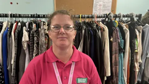 Big Venture Centre stepping in where 'everyone is struggling' BBC Jane Platt in a bright pink polo shirt. It says Big Venture Centre. Her lanyard says the same. She has glasses and is smiling. There are lots of clothes on a big rack behind her.