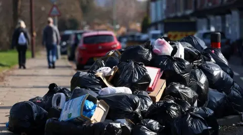 Birmingham bin strike could last until December after union vote EPA A large pile of black bin bags, with a few carboard boxes and other bits of rubbish interspersed throughout. The pile appears to be partly on the pavement and partly on the road. Cars are parked in the background, and two people are walking on the pavement away in the distance.
