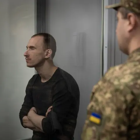 A Russian soldier takes the stand for an execution BBC Dmitriy Kurashov in the dock. He has short hair and is wearing a dark long-sleeved top. Part of a Ukrainian uniform can be seen in front of him.
