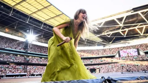 Ticket touts employing workers to bulk-buy for concerts Getty Images Singer Taylor Swift in a long green dress holding a microphone crouching down smiling while on stage with the crowd behind her at Wembley Stadium in London