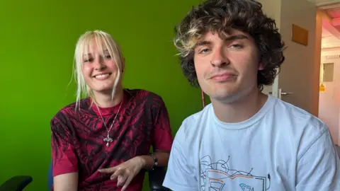 Y2K core is about nostalgia and escapism BBC A young woman with blond hair and young man with brown, curly hair sit in a room with bright green walls. The young woman wears a burgundy top with a intricate black pattern inspired by heavy metal artwork and a silver pendant on a long chain.