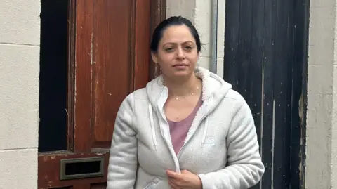 Police deploy more officers after 'mob violence' Mila Kolev wears a grey hooded cardigan and a pink t-shirt as she stands outside her home in Ballymena. Her dark brown wooden front door has a panel missing in it. She is looking at the camera with a neutral expression on her face.
