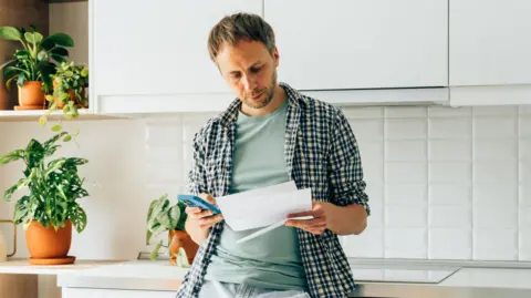 Seven ways the Spending Review affects you Getty Images Man leans against a work surface in a kitchen holding paperwork and a phone.